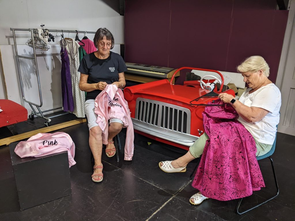 Therie and Valerie hard at work on the splendid “Pink Lady” jackets, amongst others, with the “Greased Lightnin’” car in the background.
